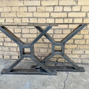 Puede incluir: Dos patas de mesa de acero en bruto con un diseño geométrico. Las patas están construidas con barras de metal soldadas, formando una base en forma de X con un detalle cuadrado en el centro. Las patas están diseñadas para soportar una mesa.