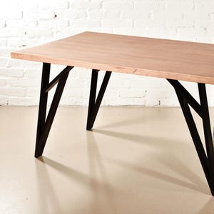 May include: A rectangular wooden table with a light brown surface and black metal legs. The legs have an angular design. The table is set against a white brick wall.