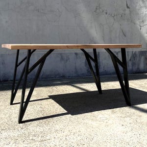 May include: A rectangular wooden table with a light brown tabletop and black metal legs. The legs have a geometric, angular design. The table is set against a concrete wall.