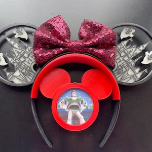 May include: Black mouse ears with a red bow and a red frame with a picture of Buzz Lightyear from Toy Story. The ears have a white silhouette of Cinderella's castle and ghosts.