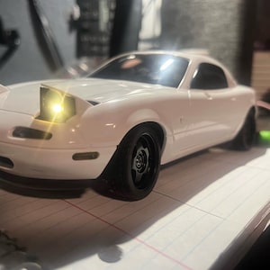 May include: A white scale model sports car featuring pop-up headlights, one of which is illuminated with a bright yellow light. The vehicle has a smooth, glossy exterior finish, black wheels, and a low-profile design with a rounded front bumper.