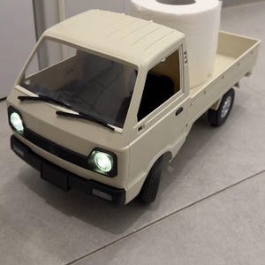 May include: A beige toy pickup truck with a large roll of white toilet paper in the bed. The truck has working headlights and a black windshield wiper. The truck is on a gray tiled floor.