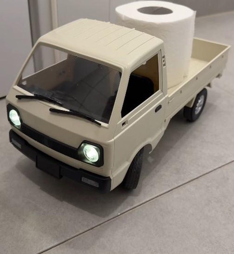 May include: A beige toy pickup truck with a large roll of white toilet paper in the bed. The truck has working headlights and a black windshield wiper. The truck is on a gray tiled floor.