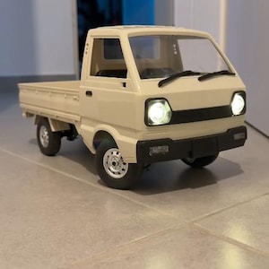 May include: A beige toy pickup truck with a black front bumper and working headlights. The truck has a flatbed and silver wheels. The headlights are illuminated, and the truck is sitting on a tiled surface.