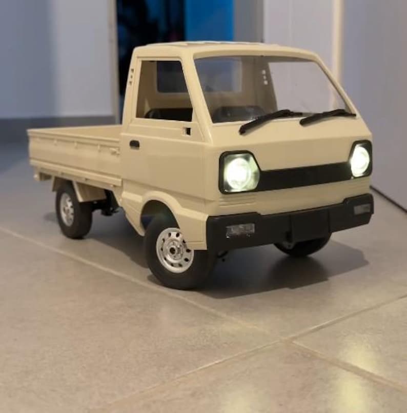 May include: A beige toy pickup truck with a black front bumper and working headlights. The truck has a flatbed and silver wheels. The headlights are illuminated, and the truck is sitting on a tiled surface.