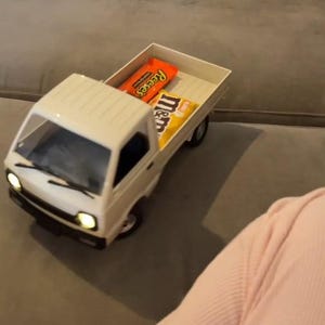 May include: A white toy pickup truck with its headlights on, carrying a Reese's candy bar and a bag of M&M's. The truck is on a gray surface.