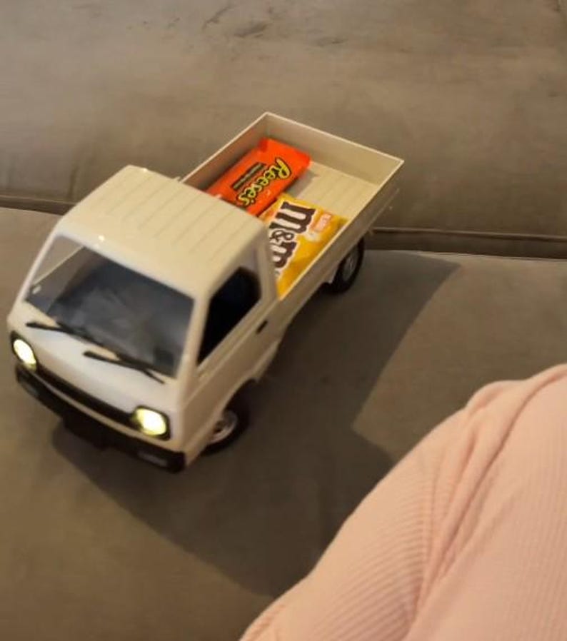 May include: A white toy pickup truck with its headlights on, carrying a Reese's candy bar and a bag of M&M's. The truck is on a gray surface.