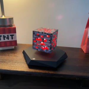 Peut inclure: Lampe cube pixélisée, ressemblant à un bloc de minerai Minecraft, flottant au-dessus d'une base hexagonale noire. Le cube est gris avec des accents rouges lumineux. Une lampe sur le thème TNT et un objet rouge sont en arrière-plan.