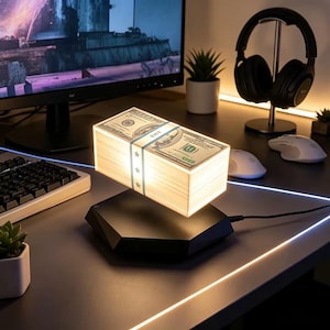 May include: A levitating rectangular lamp shaped like a stack of one hundred dollar bills. The lamp features a black hexagonal base and emits a warm white glow from the sides of the currency stack, which is secured by a blue paper band.