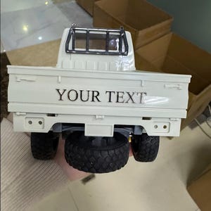 May include: A white toy pickup truck with black tires and a black roll bar. The truck bed has the text "YOUR TEXT" in silver lettering. The truck is a miniature replica.