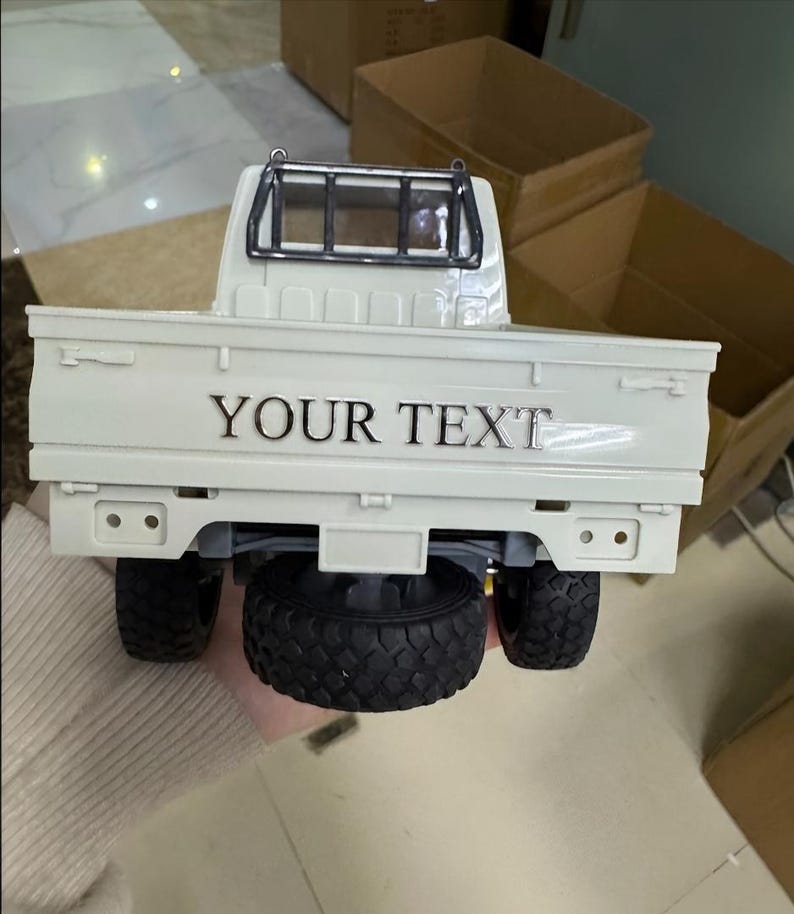 May include: A white toy pickup truck with black tires and a black roll bar. The truck bed has the text "YOUR TEXT" in silver lettering. The truck is a miniature replica.