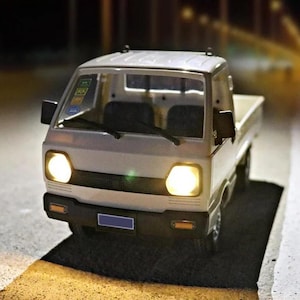 May include: A white toy pickup truck with illuminated headlights and taillights. The miniature truck has a detailed design, including a windshield, side mirrors, and a cargo bed. The truck is parked on a dark surface, with the headlights casting a warm glow.
