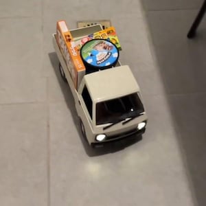 May include: A miniature white pickup truck toy with a cargo bed filled with boxes and a round disc. The truck has headlights and a windshield. The items in the bed appear to be packaged goods.