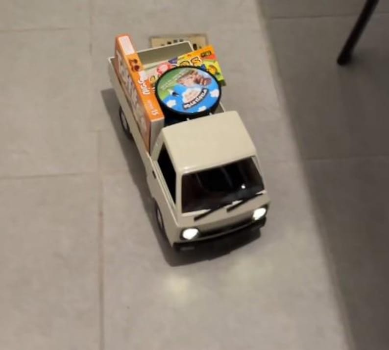 May include: A miniature white pickup truck toy with a cargo bed filled with boxes and a round disc. The truck has headlights and a windshield. The items in the bed appear to be packaged goods.