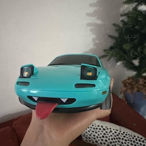 Pode incluir: Um carro de brinquedo azul turquesa com uma cara de desenho animado, incluindo uma língua vermelha que se projeta. O carro tem faróis pretos e um pára-choques preto.
