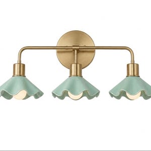 May include: A three-light wall fitting featuring a brushed brass horizontal bar and circular wall mount. Three light sockets are attached to the bar, each holding a pale mint green ceramic shade with a ruffled, scalloped edge and a light bulb.
