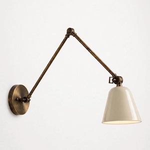 Può includere: Una lampada da parete con finitura in ottone anticato sulla base circolare e sul braccio articolato regolabile in due parti. La lampada include un paralume liscio a forma di campana color bianco sporco che dirige la luce verso il basso.