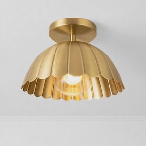 May include: A flush mount ceiling light fitting featuring a brushed brass finish. The dome-shaped shade has a scalloped edge design and a central clear glass bulb visible underneath. The fitting attaches to the ceiling with a circular brass canopy.
