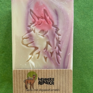 May include: A rectangular bar of soap with a carved alpaca design and pink and purple swirls. The soap is wrapped in clear plastic and has a brown label with the text "Heuberg Alpaka" and "Heckenrose".