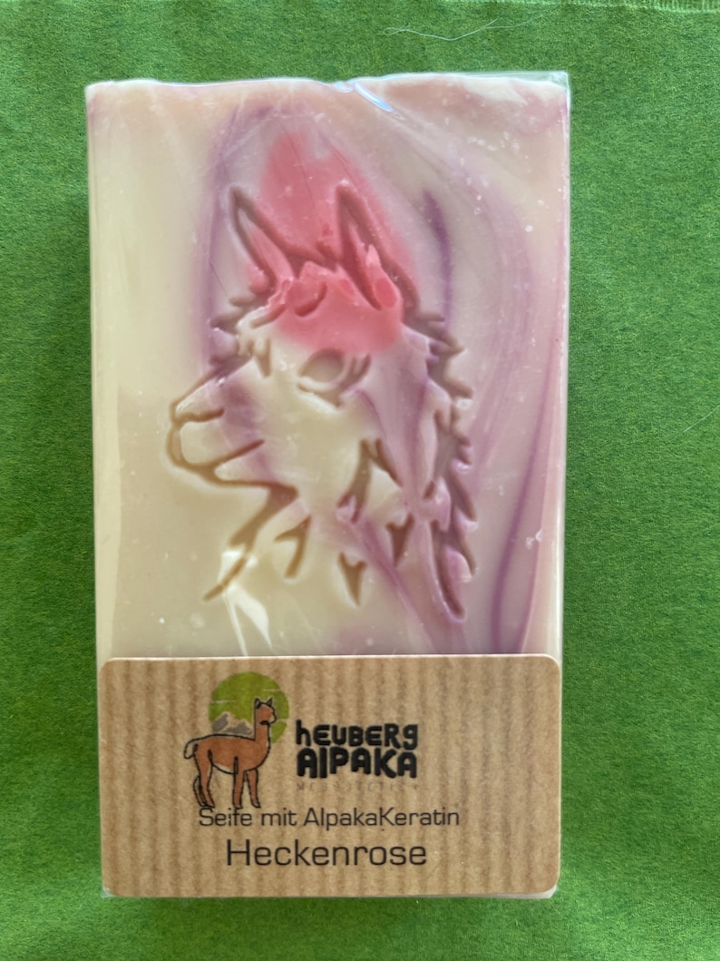 May include: A rectangular bar of soap with a carved alpaca design and pink and purple swirls. The soap is wrapped in clear plastic and has a brown label with the text "Heuberg Alpaka" and "Heckenrose".