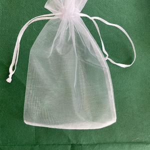 May include: A small, white, sheer organza drawstring bag. The bag is made of a delicate, see-through fabric and features a gathered top with a white drawstring closure. The bag is empty and sits on a green surface.