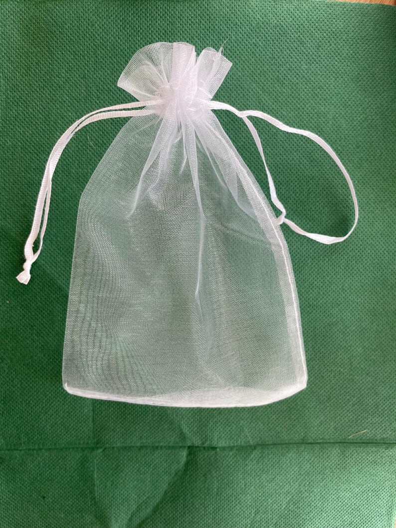 May include: A small, white, sheer organza drawstring bag. The bag is made of a delicate, see-through fabric and features a gathered top with a white drawstring closure. The bag is empty and sits on a green surface.