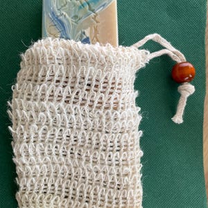 May include: A bar of soap with blue and white swirls peeks out of a cream-colored woven soap saver bag. The bag has a drawstring closure with a small orange bead. The background is a dark green fabric.