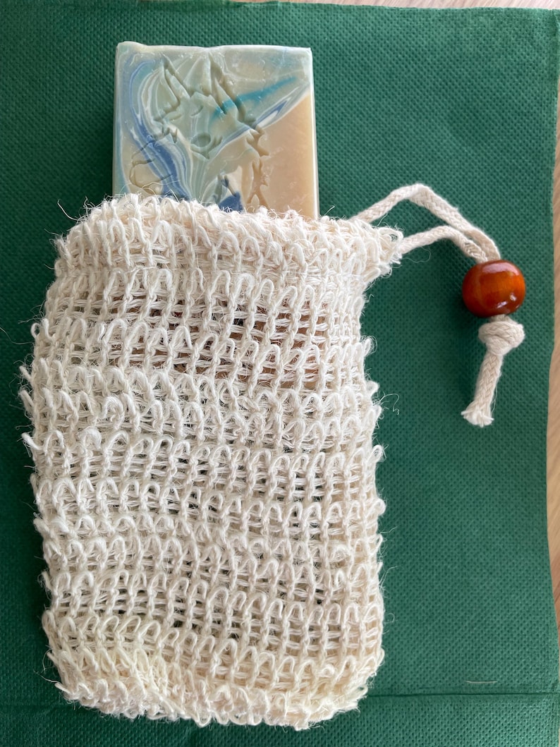 May include: A bar of soap with blue and white swirls peeks out of a cream-colored woven soap saver bag. The bag has a drawstring closure with a small orange bead. The background is a dark green fabric.