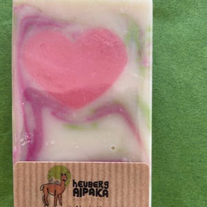 May include: A rectangular bar of soap with a pink heart design in the center, swirled with white, pink, and green hues. The soap has a brown label with an alpaca illustration and text in German.