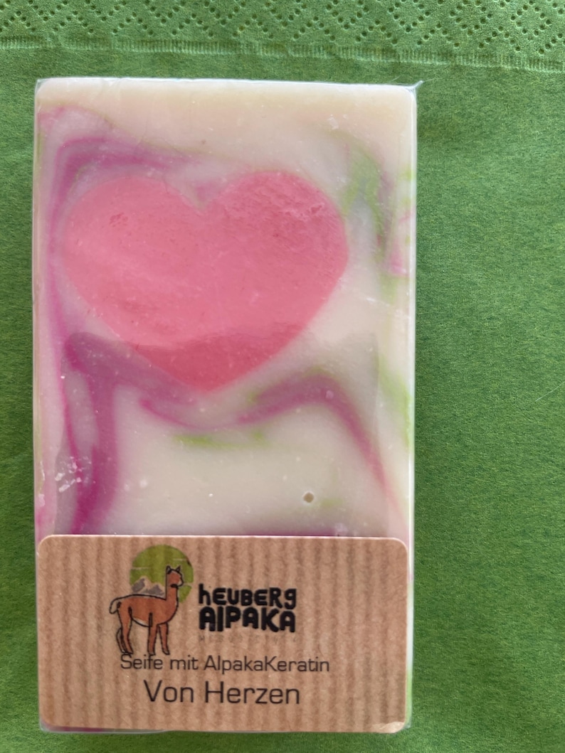 May include: A rectangular bar of soap with a pink heart design in the center, swirled with white, pink, and green hues. The soap has a brown label with an alpaca illustration and text in German.