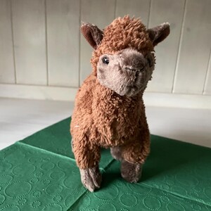 May include: A brown alpaca soft toy with a fluffy body and detailed facial features. The alpaca stands on a green patterned surface. The toy has small ears and a soft, plush texture.