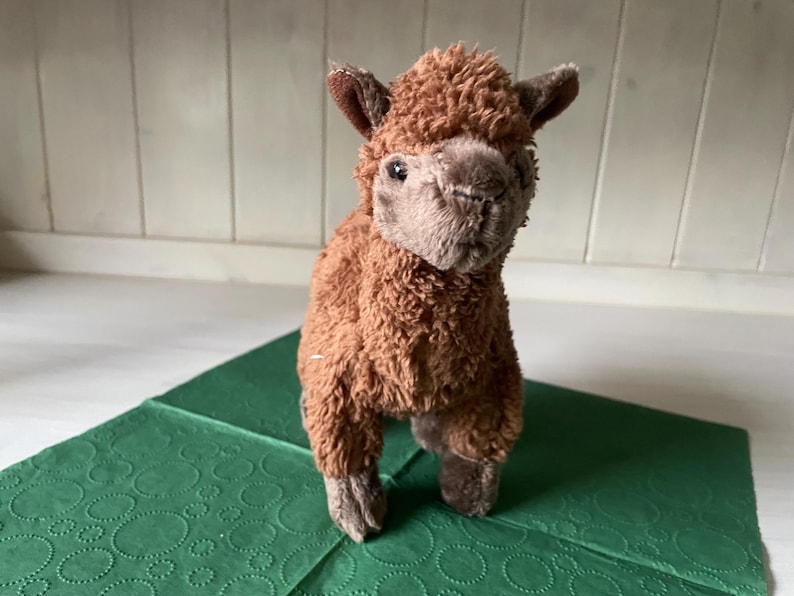 May include: A brown alpaca soft toy with a fluffy body and detailed facial features. The alpaca stands on a green patterned surface. The toy has small ears and a soft, plush texture.