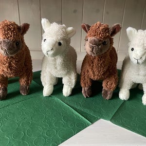 May include: Four soft toy lambs, two brown and two white, are displayed on a green patterned surface. The lambs have detailed features, including small ears and expressive faces. These cuddly toys would be ideal for children.
