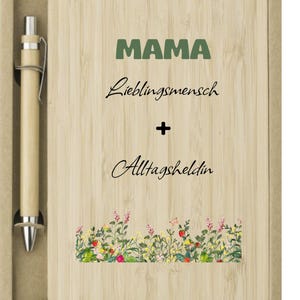 May include: A wooden notepad and pen set. The notepad has the word "MAMA" in green, with "Lieblingsmensch + Alltagsheldin" written below. A floral design is at the bottom. The pen is light brown.