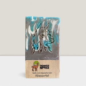 May include: A rectangular bar of soap with a gray base and a blue and white marbled top. An embossed image of an alpaca is in the center. The bottom of the soap has a brown label with the text "Heuberg Alpaka" and "Wasserfall".