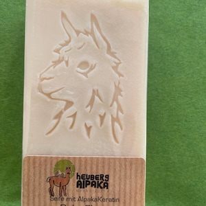 May include: A rectangular bar of soap with an embossed alpaca design. The soap is light beige and wrapped in clear plastic. A brown label at the bottom reads "heuberg alpaka" and "Baby Shea". The background is green.