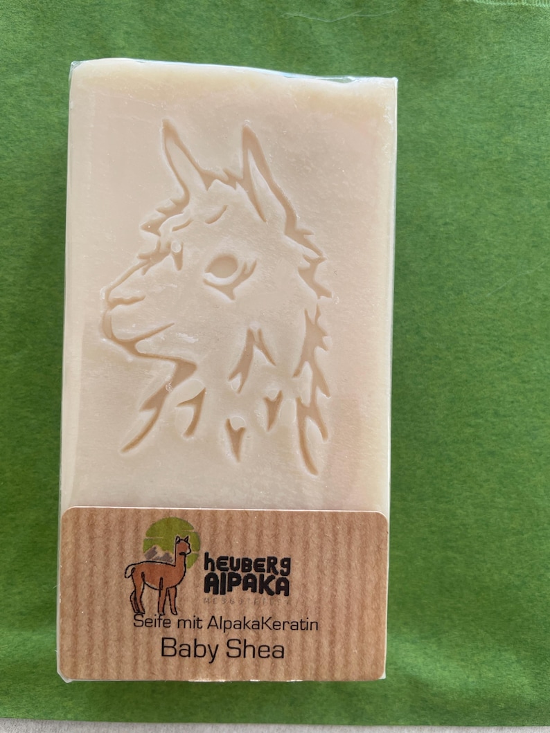 May include: A rectangular bar of soap with an embossed alpaca design. The soap is light beige and wrapped in clear plastic. A brown label at the bottom reads "heuberg alpaka" and "Baby Shea". The background is green.