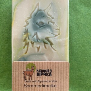 May include: A rectangular bar of soap with a carved alpaca design in shades of green and white. The soap is wrapped in clear plastic and has a brown label with the text "heuberg AIPAKA" and "Seife mit Alpakakeratin Sommerlimette".