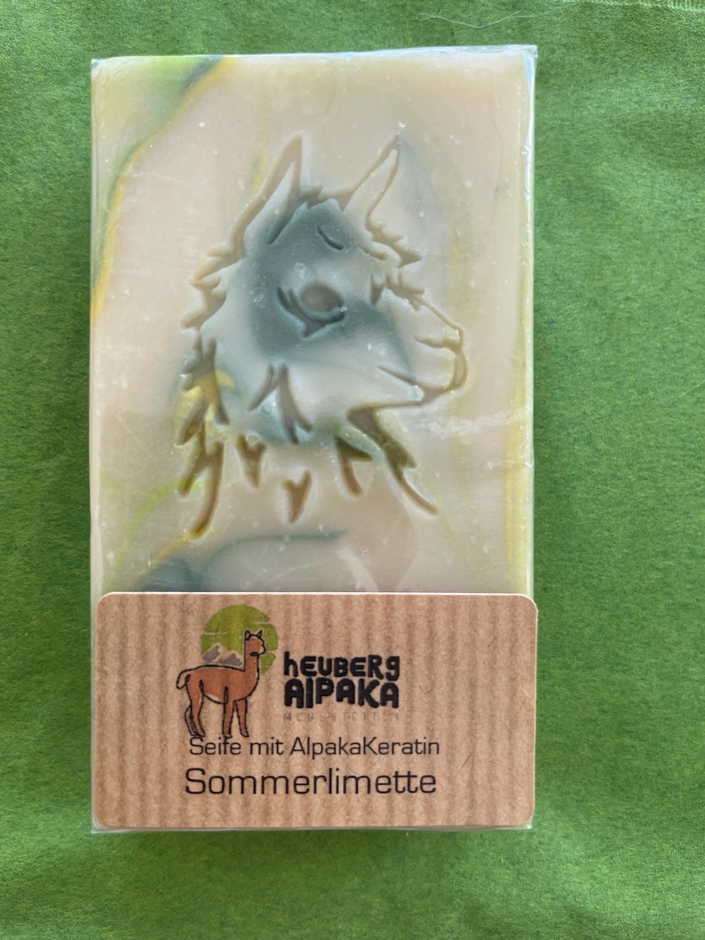 May include: A rectangular bar of soap with a carved alpaca design in shades of green and white. The soap is wrapped in clear plastic and has a brown label with the text "heuberg AIPAKA" and "Seife mit Alpakakeratin Sommerlimette".