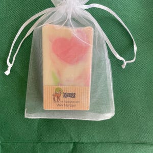 May include: A bar of soap with a pink heart design is presented in a white organza bag. The soap has a light beige base with pink swirls. A small label with text and a llama graphic is visible on the soap.