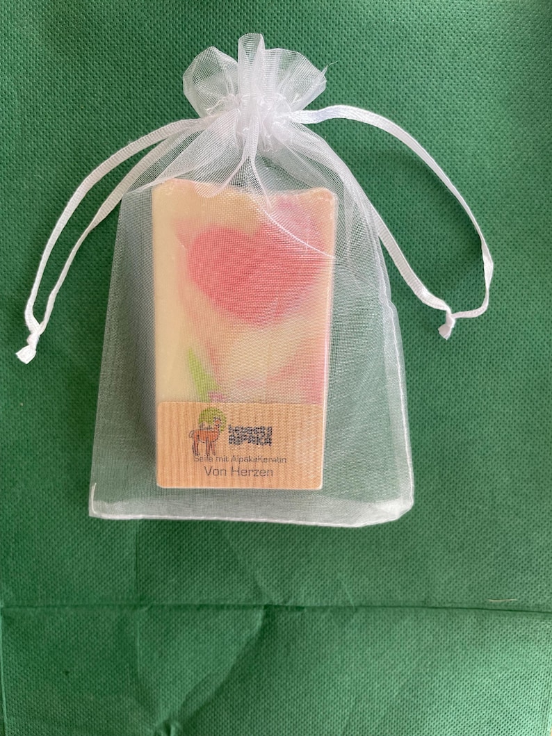 May include: A bar of soap with a pink heart design is presented in a white organza bag. The soap has a light beige base with pink swirls. A small label with text and a llama graphic is visible on the soap.