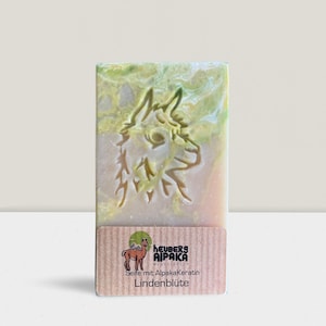 May include: A rectangular bar of soap with a carved alpaca design. The soap has a marbled appearance with green and yellow hues. A brown label at the bottom features an alpaca illustration and text.