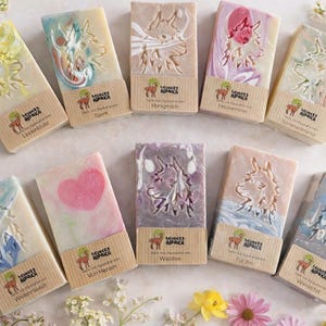 May include: An assortment of handmade soaps with various designs and colors. Each rectangular bar features a unique embossed pattern, including floral and animal motifs. The soaps are arranged with floral accents and small tags with text.