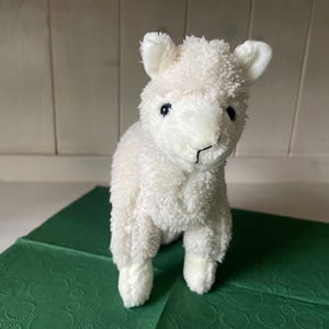 May include: A white plush llama toy with a fluffy texture, small ears, and black eyes. The soft toy is placed on a green surface. The llama is a soft toy.