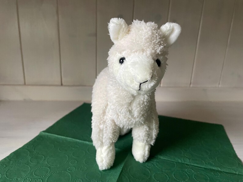 May include: A white plush llama toy with a fluffy texture, small ears, and black eyes. The soft toy is placed on a green surface. The llama is a soft toy.