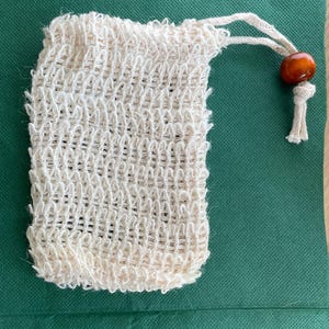 May include: A natural-colored, woven soap bag with a drawstring closure and a wooden bead. The bag is made of a textured, open-weave material. The drawstring is made of a matching natural fiber.