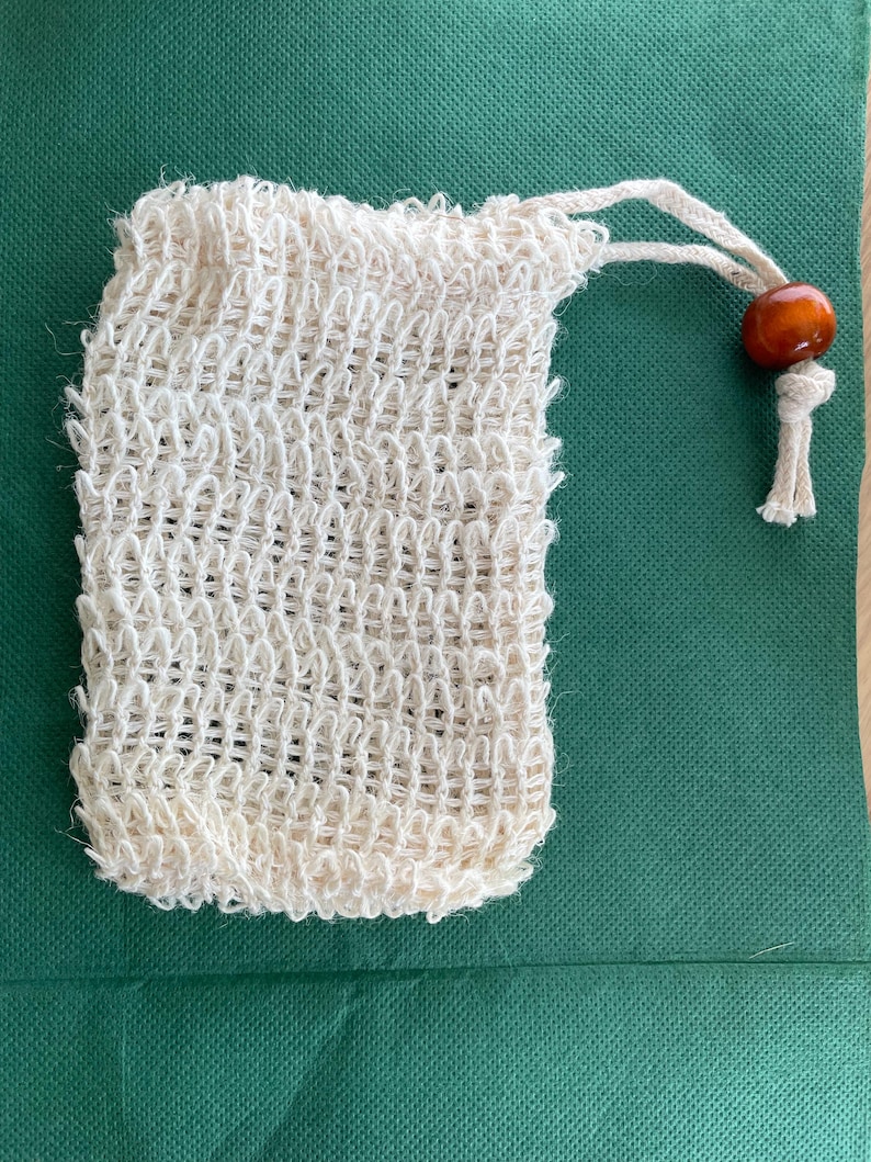 May include: A natural-colored, woven soap bag with a drawstring closure and a wooden bead. The bag is made of a textured, open-weave material. The drawstring is made of a matching natural fiber.