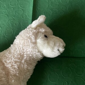 May include: A soft, white llama toy with a textured, fluffy coat. The llama has small, dark eyes and a detailed snout. The toy is set against a green background with a circular pattern.