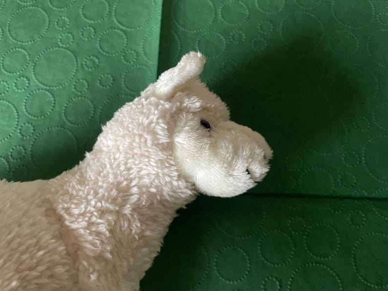 May include: A soft, white llama toy with a textured, fluffy coat. The llama has small, dark eyes and a detailed snout. The toy is set against a green background with a circular pattern.