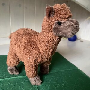 May include: A brown plush llama toy with a fluffy body, standing on a green surface. The llama has a detailed face with dark eyes and a soft, textured appearance. The toy is a cute and cuddly item.